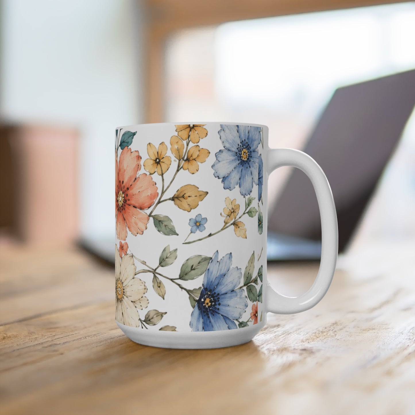 A white ceramic mug with a colorful watercolor floral pattern, sitting on a wooden table with a blurred laptop in the background.