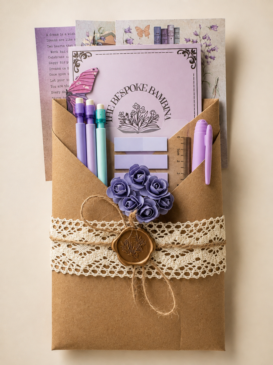 A brown kraft paper envelope-style organizer filled with stationery, tied with a cream lace ribbon and a wax seal. Visible contents include purple and pastel pens, a lavender card reading 'The Lakespoe Bakery,' decorative paper, and small purple paper flowers.