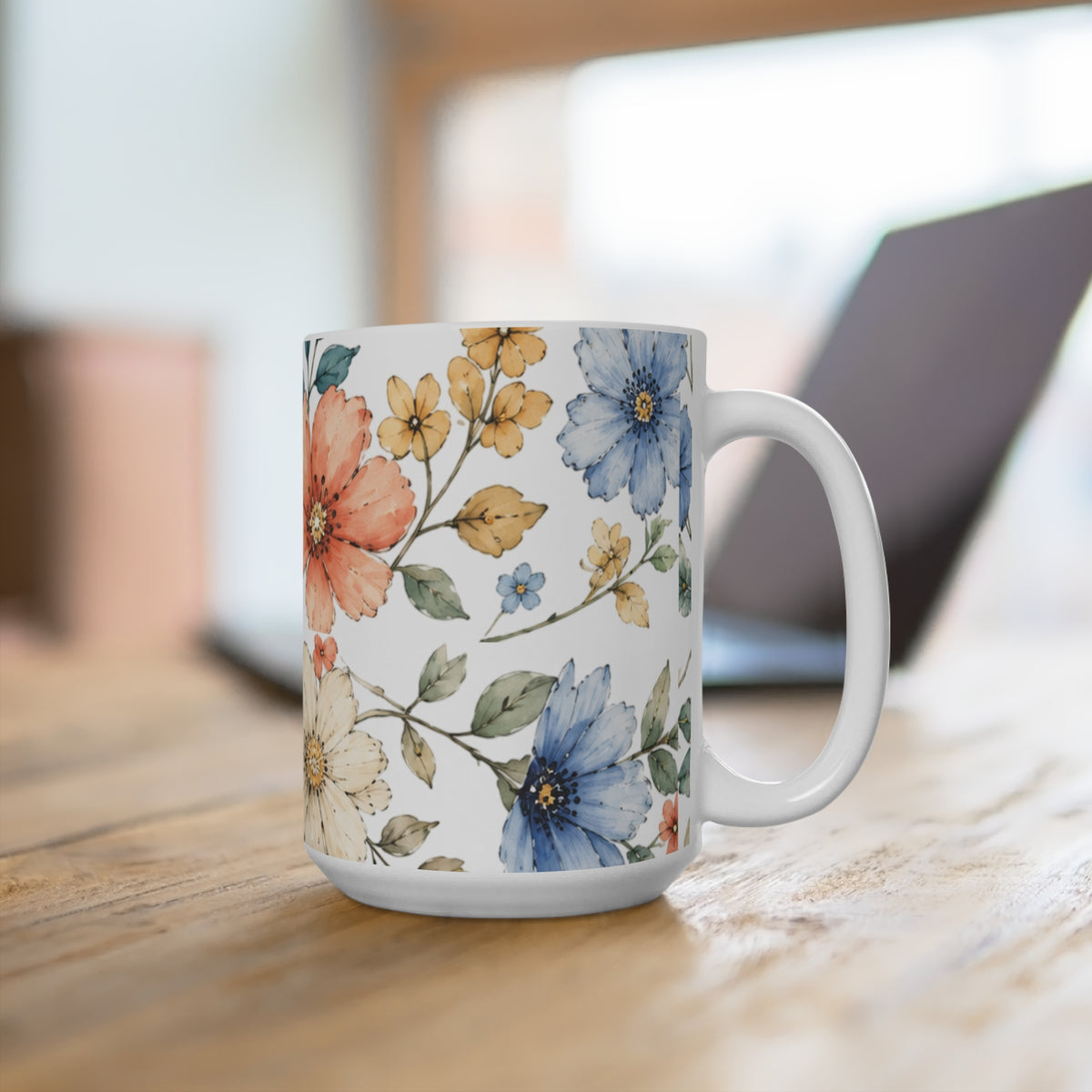A white ceramic mug with a colorful watercolor floral pattern, sitting on a wooden table with a blurred laptop in the background.