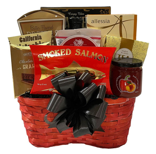 A red woven gift basket filled with assorted gourmet snacks and tea, featuring a large black ribbon bow on the front. Visible items include a red package of smoked salmon, a jar of fruit preserves, chocolate-branded boxes, and allessia tea.