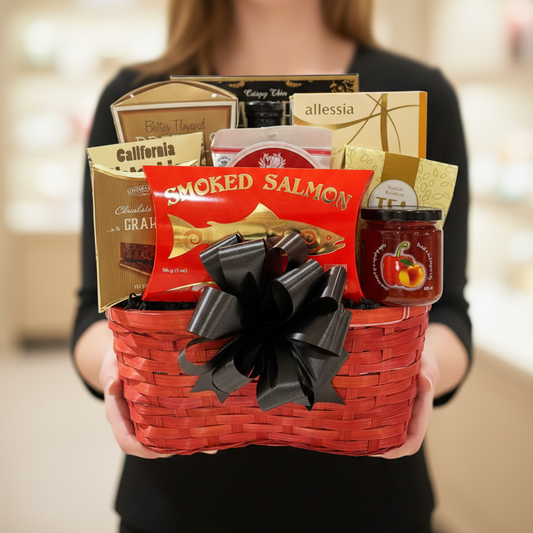 A red woven gift basket filled with assorted gourmet snacks and tea, featuring a large black ribbon bow on the front. Visible items include a red package of smoked salmon, a jar of fruit preserves, chocolate-branded boxes, and allessia tea.
