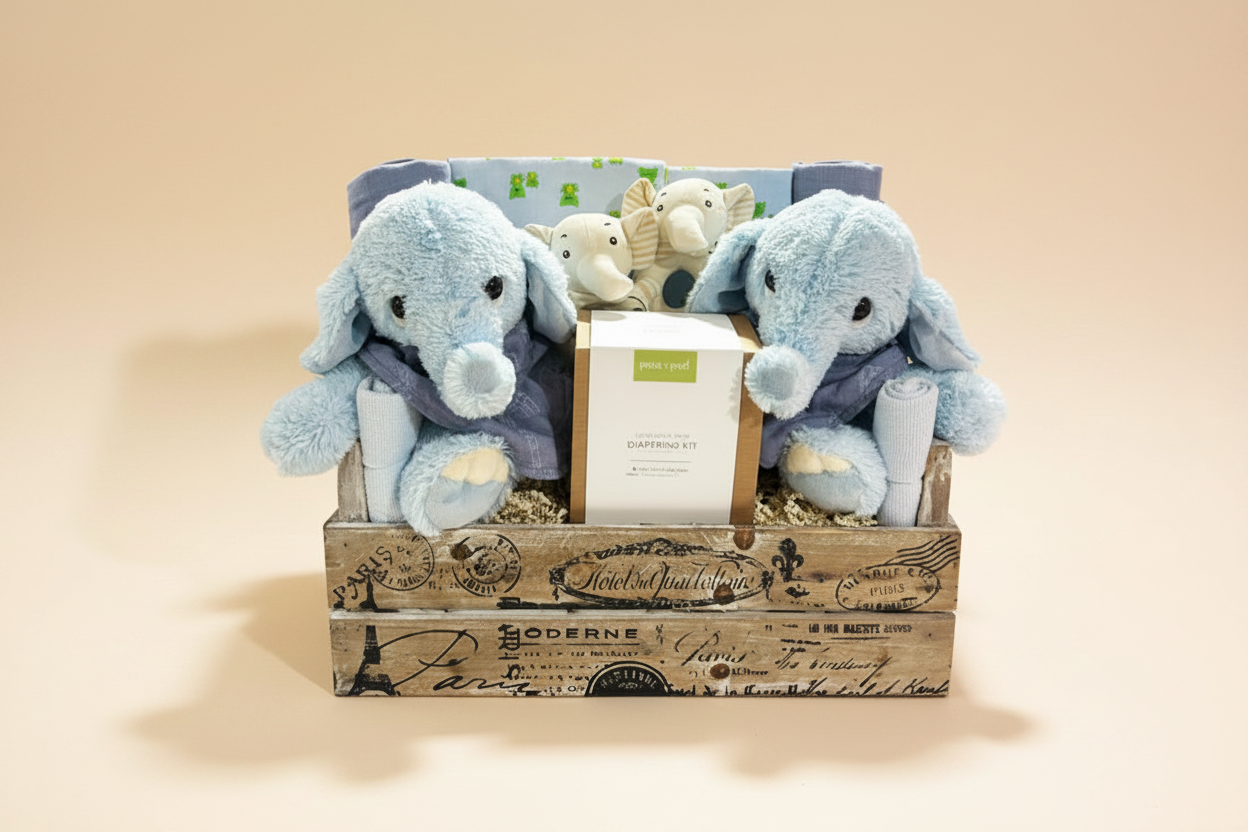 A baby gift basket set in a rustic wooden crate, featuring two light blue plush elephant toys, a small elephant plush, a box of paper napkins, a blue silicone teether, and baby accessories.
