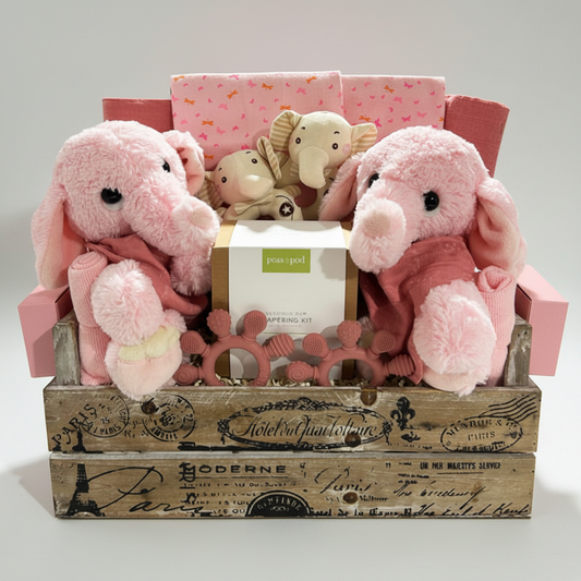 A baby gift basket set in a rustic wooden crate, featuring two pink plush elephant toys, a pink patterned fabric item, a small elephant-shaped toy, and a boxed baby gift kit labeled 'bean pod'.
