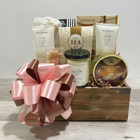 A curated spa gift basket arranged in a wooden crate with a large pink satin bow. Visible items include a jar candle, a tube of body wash, a boxed tea, a bath mat, and a round tin of candy canes, with allessia branding visible on some packages.