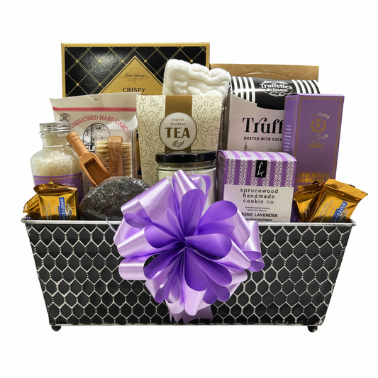 A curated gift basket filled with assorted packaged snacks, tea, soap, and small treats, arranged in a black-and-white patterned basket with a large purple bow in front. Visible packaging includes Truff, Handcrafted Homemade Soap Co.