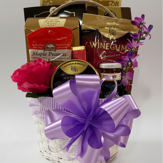 A gourmet gift basket filled with assorted snacks and treats, decorated with a large purple ribbon bow and pink flowers, set against a plain white background.