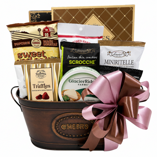 A gourmet gift basket in a dark brown metal tub with a pink and brown ribbon, filled with assorted snacks and treats including popcorn, cookies, crackers, truffles, and cheese. The basket is set against a clean white background.