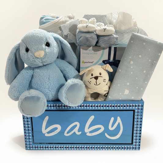 A baby gift basket filled with soft toys and baby accessories, including a large light blue plush elephant, a small dog plush, a gray blanket with white dots, striped baby socks, and a blue basket with the word "baby" on the front.