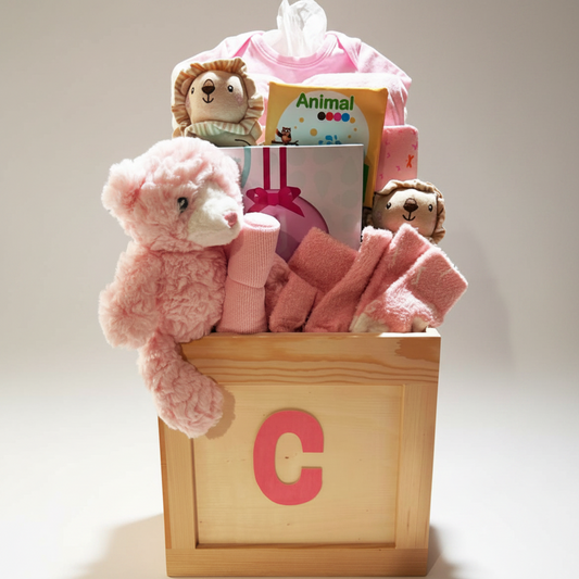 A baby gift basket arranged in a wooden crate with a large pink letter C, filled with pink baby clothes, a plush teddy bear, pink socks, and small stuffed lion toys on a white background.