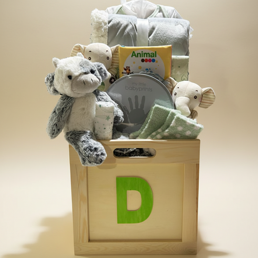 A baby gift basket arranged in a wooden storage box with a large green letter D on the front. The basket contains a plush elephant toy, folded blankets, a baby book, a tin of baby fingerprints, and a small cup, all photographed on a white background.