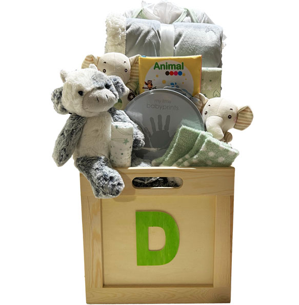 A baby gift basket arranged in a wooden storage box with a large green letter D on the front. The basket contains a plush elephant toy, folded blankets, a baby book, a tin of baby fingerprints, and a small cup, all photographed on a white background.