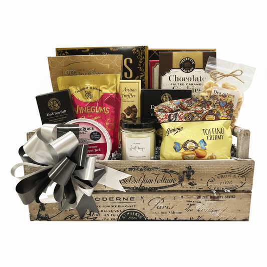 A gourmet gift basket filled with assorted snacks and treats, including a bag of winegums, chocolate salted caramel cookies, toffino creamer, and a jar of salted caramel, arranged in a rustic wooden crate with a large silver and black ribbon bow.