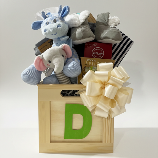 A baby gift basket in a wooden crate with a large decorative bow, filled with plush toys, baby shoes, and boxed items. The crate has a green letter D on the front.