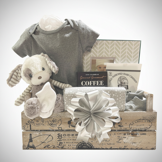 A baby gift basket set in a rustic wooden crate with a large white bow, featuring a gray short-sleeve baby shirt, a plush stuffed bunny, a box of coffee, and baby accessories on a white background.