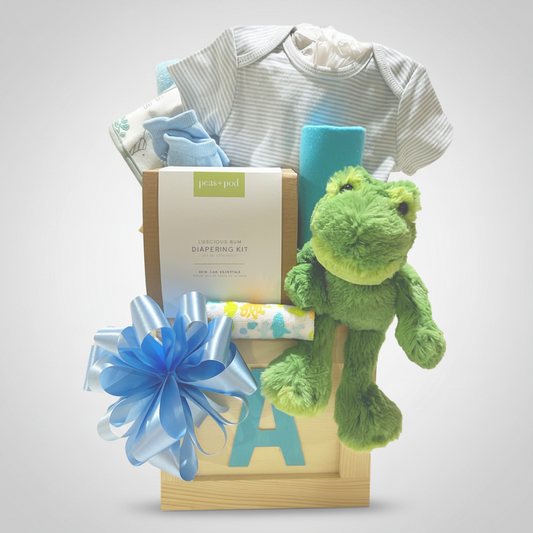 A baby gift basket set from Jessup Dot featuring a light blue and white striped baby onesie, a green plush frog toy, a blue and white diaper kit box, a blue ribbon bow, and a wooden letter A tray, all arranged on a white background.