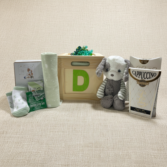 A baby gift basket set featuring a wooden crate with a green letter D, a plush gray and white puppy toy, a roll of light green baby blankets, packaged baby socks, a box of Cappuccino espresso powder, and a white box with baby illustrations, all arranged on a white background.