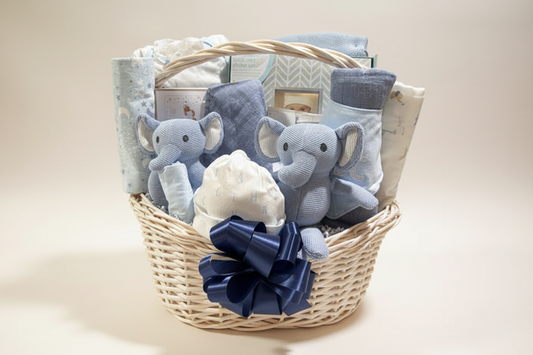 A wicker gift basket filled with baby essentials, including two light blue plush elephant toys, folded blankets, a white bonnet, a navy ribbon bow, and boxed items, photographed on a plain white background.