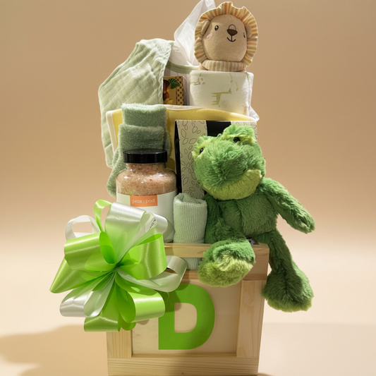 A baby gift basket arranged in a wooden crate with a large green and white bow, featuring a plush green frog toy, a lion plush, a folded towel, a jar of bath salts, and baby wipes on a white background.