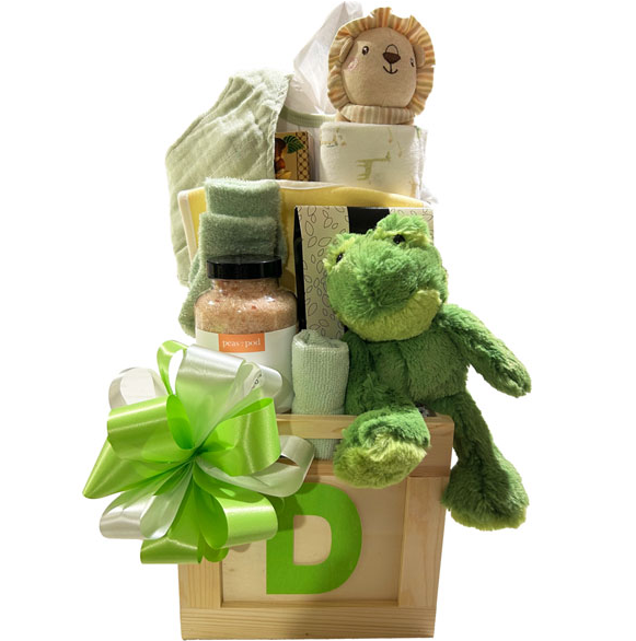 A baby gift basket arranged in a wooden crate with a large green and white bow, featuring a plush green frog toy, a lion plush, a folded towel, a jar of bath salts, and baby wipes on a white background.