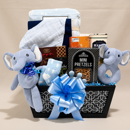 A baby gift basket set against a white background, featuring a black basket with a white geometric pattern and a large blue satin bow. Inside the basket are a light blue and white swaddle blanket, a light blue plush elephant toy with a blue bow on the left, and a matching light blue plush elephant toy on the right.