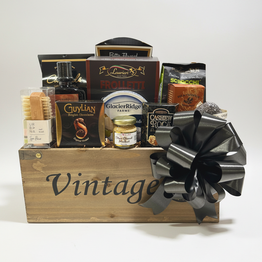 A wooden gift basket filled with assorted gourmet snacks, crackers, and packaged food items, decorated with a large black ribbon bow. Visible brands include Vintage on the basket, Glacier Ridge Farms on the cheese jar, and Cashewy Crocks on the crackers.