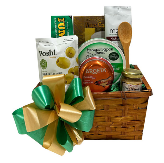 A gourmet gift basket filled with various food items and a large green and gold bow. The basket contains Poshi green olives, Glacier Ridge Farms salmon tartare, Argeta salmon tartare, a jar of honey, a wooden spoon, and other packaged snacks, all arranged in a brown woven basket.
