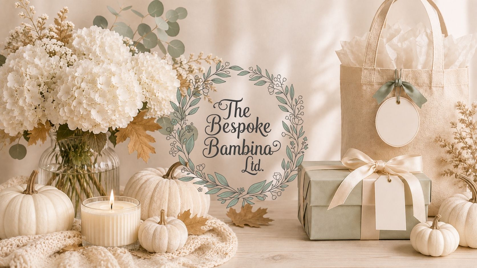 A styled autumn-themed tabletop scene with white flowers in a glass vase, small white pumpkins, a lit candle, a beige tote bag with a blank hanging tag, a wrapped gift box with a cream ribbon, and a central circular sign reading 'The Bespoke Bambina Ltd.'.