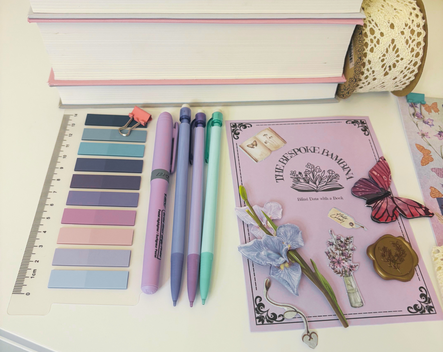 A stationery and craft supplies flat lay on a white desk, including a stack of books, a lace ribbon roll, a ruler, color swatches, pastel pens, a decorative card reading 'The Postage Paper,' and butterfly-themed paper accents.