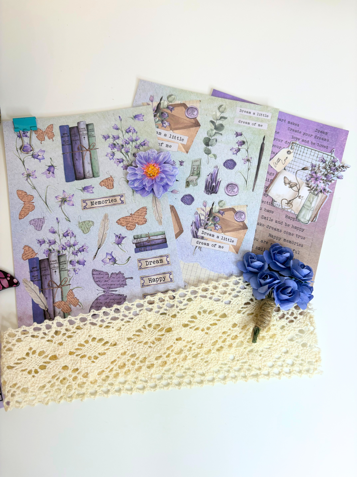 A set of decorative scrapbooking or journaling papers with vintage-style illustrations, including butterflies, books, flowers, and labels, arranged on a white surface with a cream lace runner and blue fabric flowers.